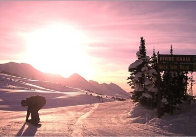 Banff Sunshine Village'da şafak vakti, güneş dağların üzerinden yükselirken pistte bir snowboardcu ve Divide Chair tabelası