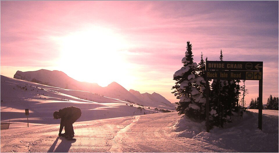 Banff Sunshine Village'da şafak vakti, güneş dağların üzerinden yükselirken pistte bir snowboardcu ve Divide Chair tabelası
