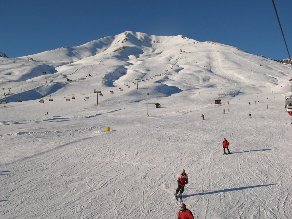 Passo del Tonale'de güneşli bir günde geniş pistte kayak yapanlar ve arkada telesiyejlerin çıktığı karlı dağ